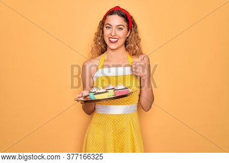 Beautiful pin-up woman with blue eyes holding chocolate cupcakes over yellow background happy with big smile doing ok sign, thumb up with fingers, excellent sign