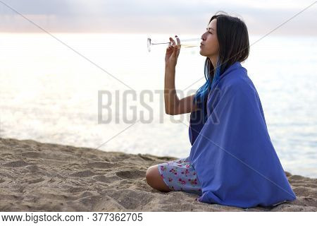 Young Girl Drinking Champagne On The Beach At Sunrise. Woman Wrapped In A Blanket Sitting On The San