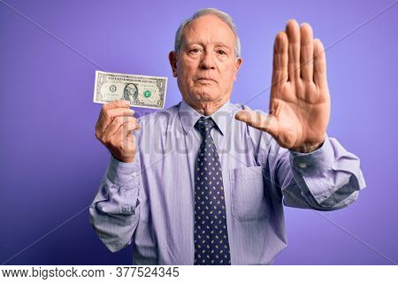 Senior grey haired man holding one dollar bank note over purple background with open hand doing stop sign with serious and confident expression, defense gesture