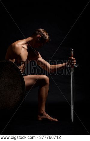 Side View Of Young Strong Warrior Standing On Knee And Looking Down. Muscular Half Naked Athlete Pos