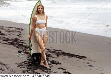Healthy Smiling Fit Woman In Bikini Stands With Surfboard, On Black Beach By The Ocean. Extrime Wate