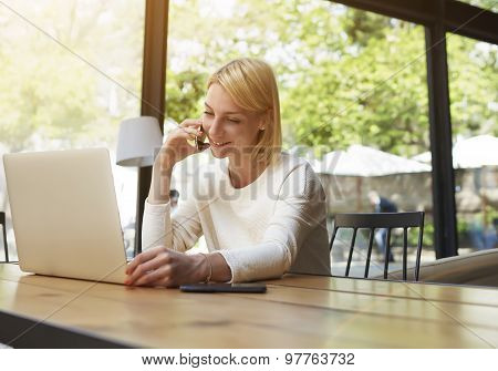 Enamored female speaking on her mobile phone with lovely smile while sitting at wooden table