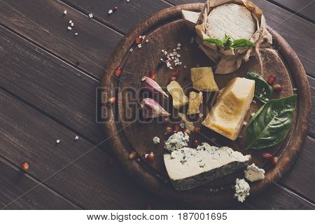 Cheese delikatessen top view on rustic wood. Wooden round desk with blue cheese roquefort, parmesan, camembert and brie cuts decorated with garlic, pomegranate and basil, french cuisine ingredients