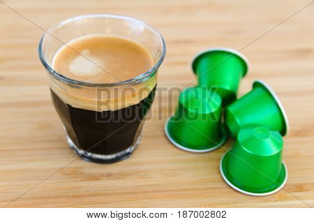 Close up of tasty espresso with foam and green coffee capsules on wooden background.