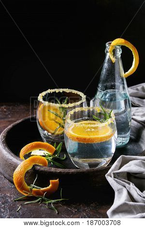 Tonic water cocktail with rosemary and orange. Two glasses and bottle with zest sugar and bubbles in terracotta tray over black texture background. Refreshing beverage alco non alcohol