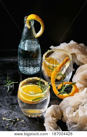 Tonic water cocktail with rosemary and orange. Two glasses and bottle with zest sugar and bubbles over black texture background with textile gauze. Refreshing beverage alco non alcohol