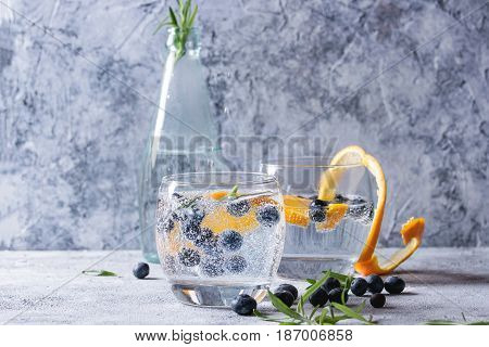 Tonic water cocktail with rosemary, blueberries and orange. Two cold glasses and bottle with zest and bubbles over gray blue texture background. Refreshing beverage alco non alcohol