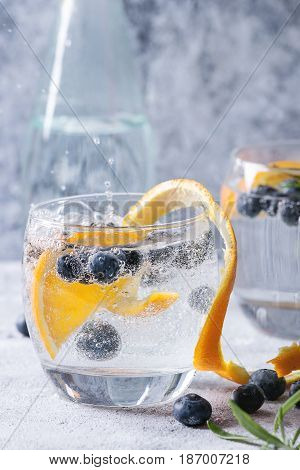 Tonic water cocktail with rosemary, blueberries and orange. Two cold glasses and bottle with zest and bubbles over gray blue texture background. Refreshing beverage alco non alcohol. Close up