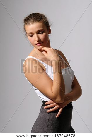 Attractive girl in white shirt with open shoulders posing in studio. Her sight is directed downward. Her arms near face and under breast. Natural color lips. Dance, art photo, ballet, fashion.