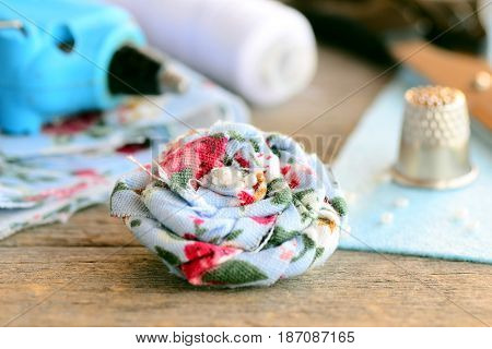 Floral linen brooch. Fabric flower brooch decorated with beads, hot glue gun, scissors, thread spool, thimble, felt sheet on vintage wooden table. Summer women brooch DIY concept. Closeup