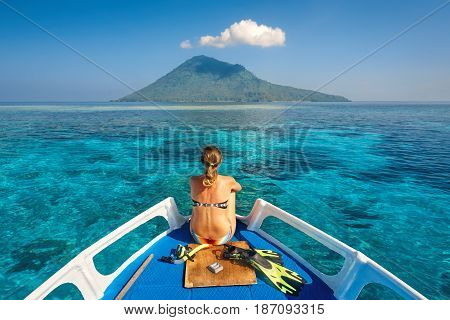 Young woman in swimsuit sit on boat with a mask and flippers looking to a clean sea and volcano Manado Tua. North Sulawesi Indonesia.