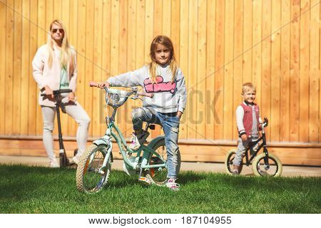 Mom with children on bicycles ride near wooden fence during day