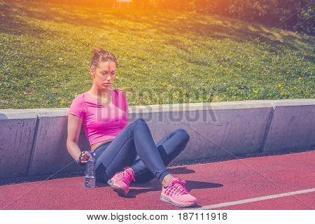 Fitness runner woman sitting on running track. Athlete girl taking a break during run to hydrate during hot summer day. Healthy active lifestyle.