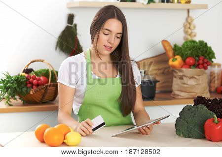 Young woman is making online shopping by tablet computer and credit card. Housewife found new recipe for cooking in a kitchen.