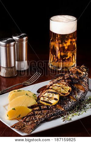 Grilled carp fish with rosemary potatoes, lemon and beer on a wooden table. Black background. Selective focus