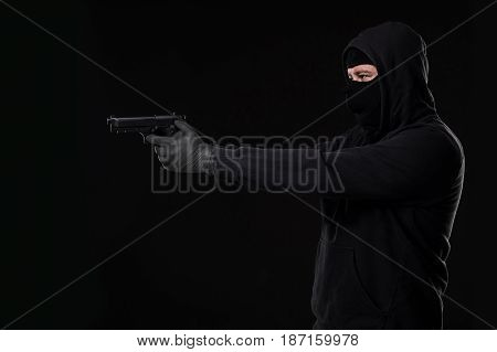 The robber in a mask with a gun pointed to the side on a black background. Man with a gun