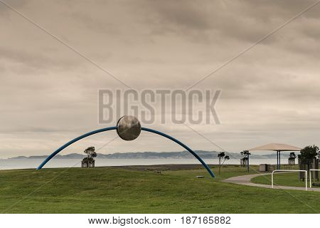 Napier New Zealand - March 9 2017: Metal Ecliptic sculpture by David Trubridge at beach park under heavy brown storm sky. Pacific Ocean and green lawn with some short trees.