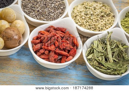 collection of superfoods in small ceramic bowls against rustic wood: macadamia nuts, hemp seed hearts, goji berry, stevia herb and chia seeds