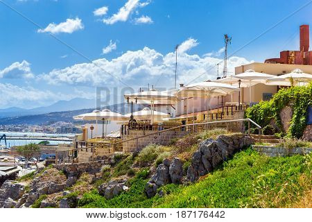 Outdoor resort cafe with white parasols on top of hill above highway along promenade Emmanouil Kefalogianni. Greek architecture on coast of Kolpos Almirou. Rethymno Crete Greece