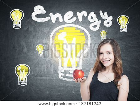 Smiling young woman in a tank top is holding a red apple and standing near a blackboard with an energy sketch drawn on it.