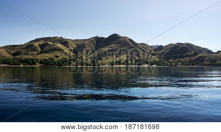 Coastline Of Mountains With Green Vegetation Reflected  In Blue Ocean Water At Labuan Bajo In Flores