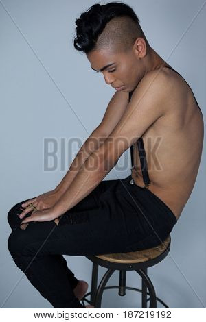 Side view of transgender wearing suspenders sitting on stool against gray background