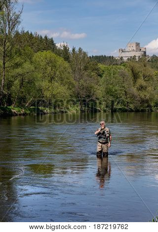 A fisherman with a pole hunts trout. A man in sport fishing tackle flies on a river. In the background on the hill is Rabi Castle. A man flying on the river Otava South Bohemia Czech republic.