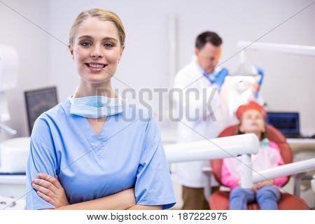 Portrait of smiling nurse with arms crossed while dentist examining patient in background