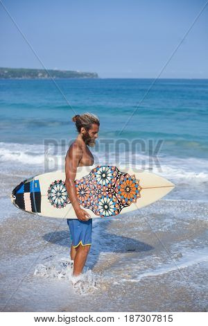 Full-length vertical portrait of an attractive bearded curly man with a surfboard is standing on beach