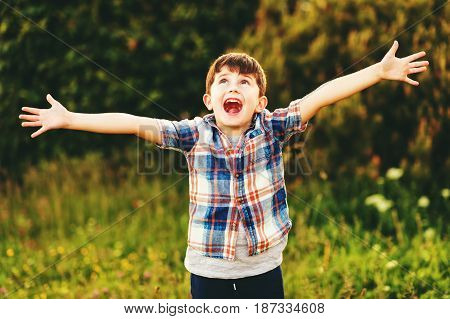 Happy kid boy of 6 year old having fun outdoors wearing blue plaid shirt, arms wide open
