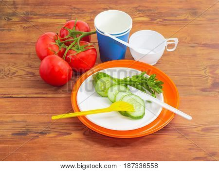 Disposable plastic plates fork knife and sliced cucumber on it claster of the tomatoes paper and plastic disposable cups with spoons beside on a surface of the old wooden planks