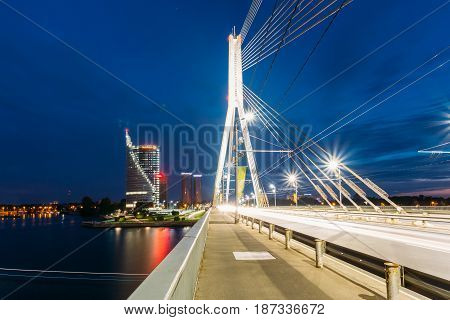 Riga, Latvia - July 01, 2016: Close View Of Vansu Cable-Stayed Bridge In Bright Night Illumination Over The Daugava River, Skyscrapers Of Downtown And Blue Sky Backgroung