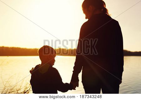 Silhouette picture of young mother and son watching sunset near water. Woman and child holding hands staying together.