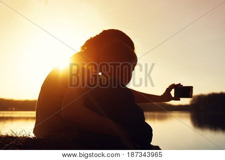 Two silhouettes of mother and her son watching sunset and making selfie picture. Young woman and child sitting on the ground near water. Happy family concept.