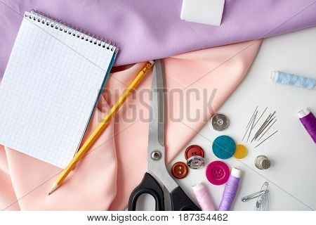 needlework and tailoring concept - scissors, sewing buttons, spools of thread, cloth and notepad with pencil