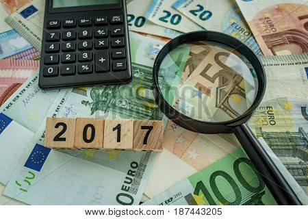 financial tax concept as magnifying glass on pile of euro banknotes calculator and number 2017 on wooden block.