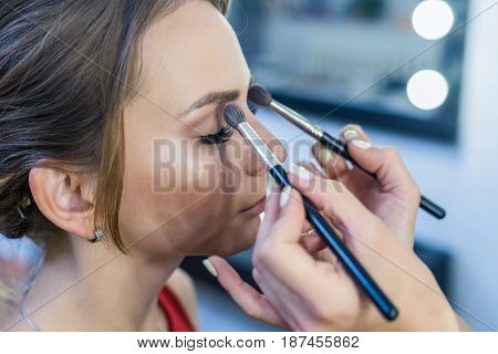 Make-up artist doing smoky eyes makeup to beautiful young girl in the studio dressing room with two brushes.