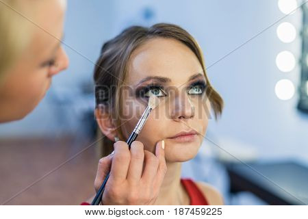 Make-up artist doing smoky eyes makeup to beautiful young girl in the studio Dressing room. The makeup artist has brush in hand