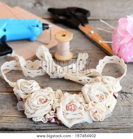 Casual floral fabric necklace, glue hot gun, scissors, thread, needle, felt on a vintage wooden table. Creating a flowers jewelry using vintage and recycled cotton fabrics, beads and felt