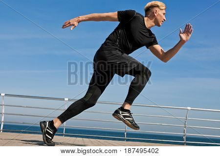Side view of man in sportswear running fast on pier with sea on background.