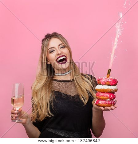 Young laughing woman holding wineglass and doughnuts with sparkler on pink background.