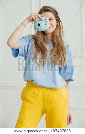 Smiling girl in casual cloth makes photo by portative camera on background of wall. Photographer young woman with long hair dressed in cosy home clothes posing in blue t-shirt and yellow trousers,