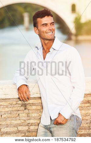 Beautiful smiling man outdoors. Tiber river from the bridge. A beautiful young Italian man posing outdoors on Milvian Bridge in the historic center of Rome, Italy. Blue shirt and jeans.