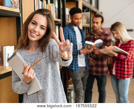 Smiling young girl student holding textbook and showing peace gesture while standing at the library with her friends on a background