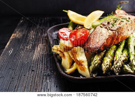 Recipe - delicious portion of grilled salmon fillet with asparagus tomatoes artichoke spices on a black wooden board. Close up copyspace selective focus.