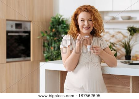 Pregnancy cravings. Positive curly-haired mom-to-be eating a healthy homemade yogurt for breakfast and smiling at the camera.
