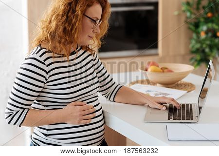 Problems to be solved. Mature woman wearing casual clothes touching a pregnant bump while working on a laptop.