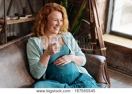 Sincere moments. Radiant pregnant woman sitting on a leather sofa with a glass of milk and smiling broadly.