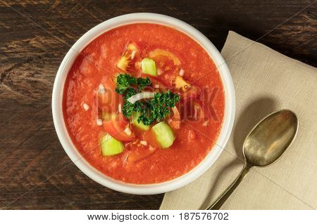 A bowl of gazpacho soup with slices of tomato, cucumber, and onion, with parsley leaves, shot from above on a dark rustic texture with a place for text