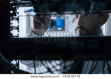 Network system. Professional handsome system administrator standing near the server rack and checking holding an internet cable while checking the network system